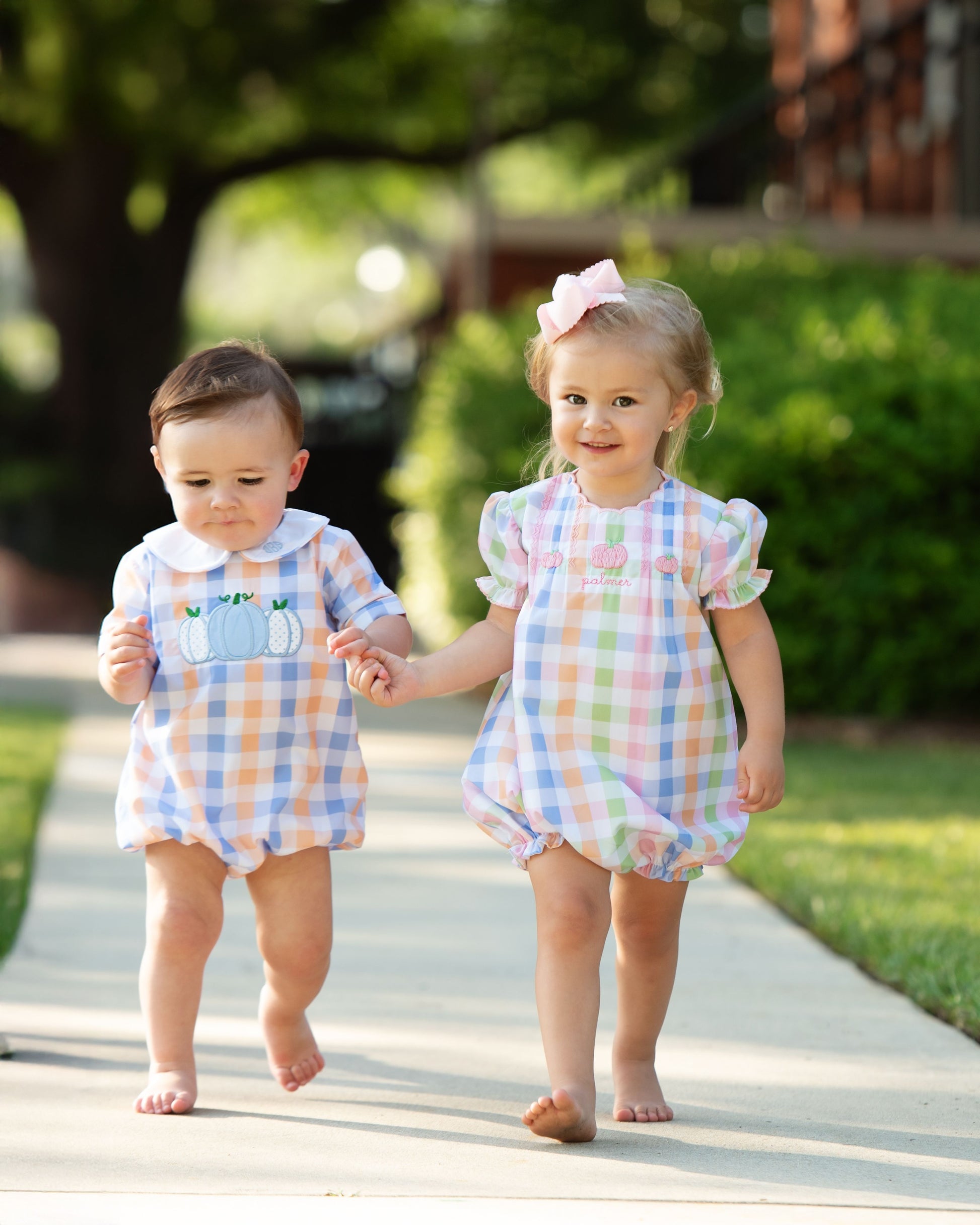 Two children in matching pumpkin outfits walking hand in hand on a sidewalk.