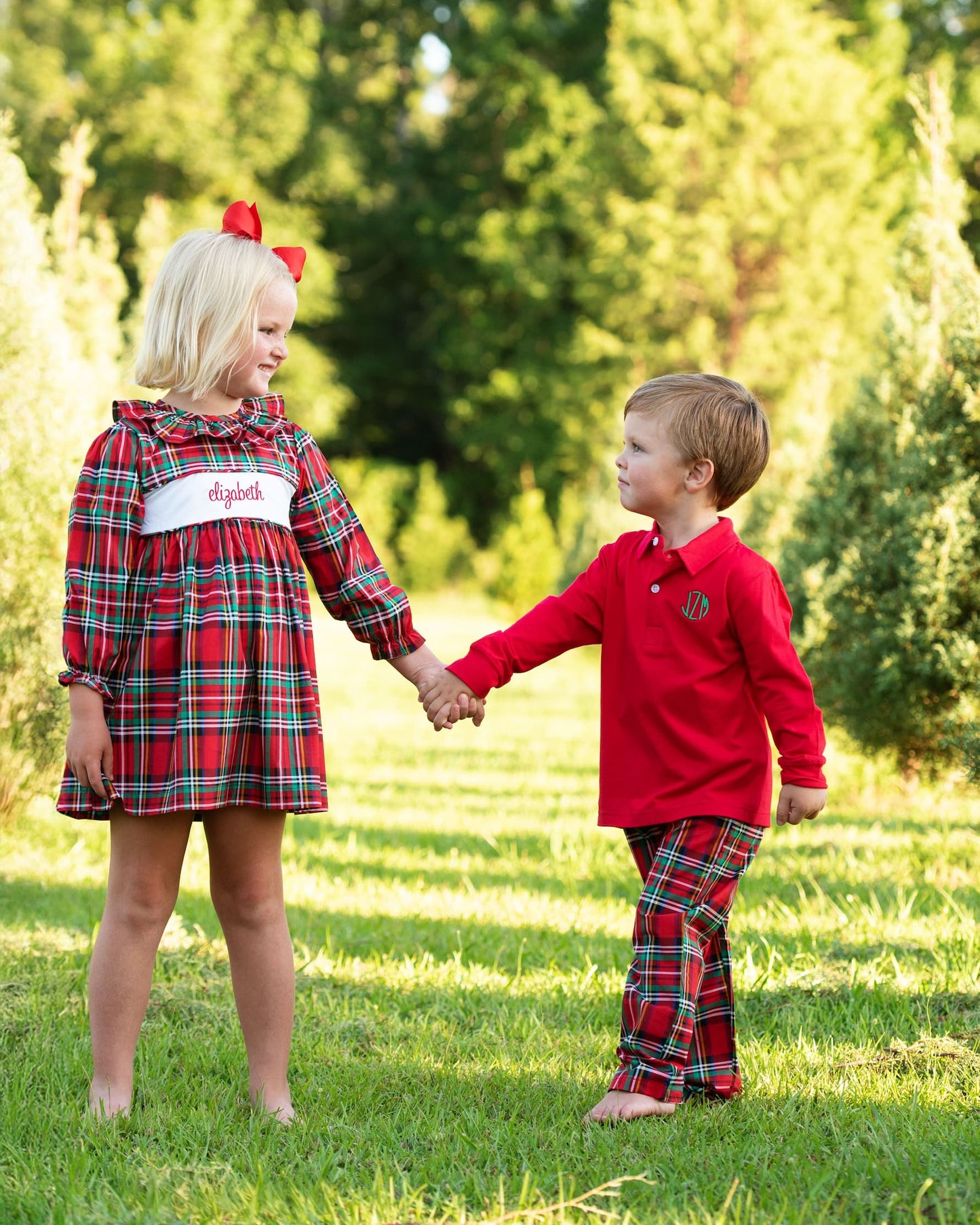 Two children in matching red plaid outfits standing in a grassy area with trees in the background.