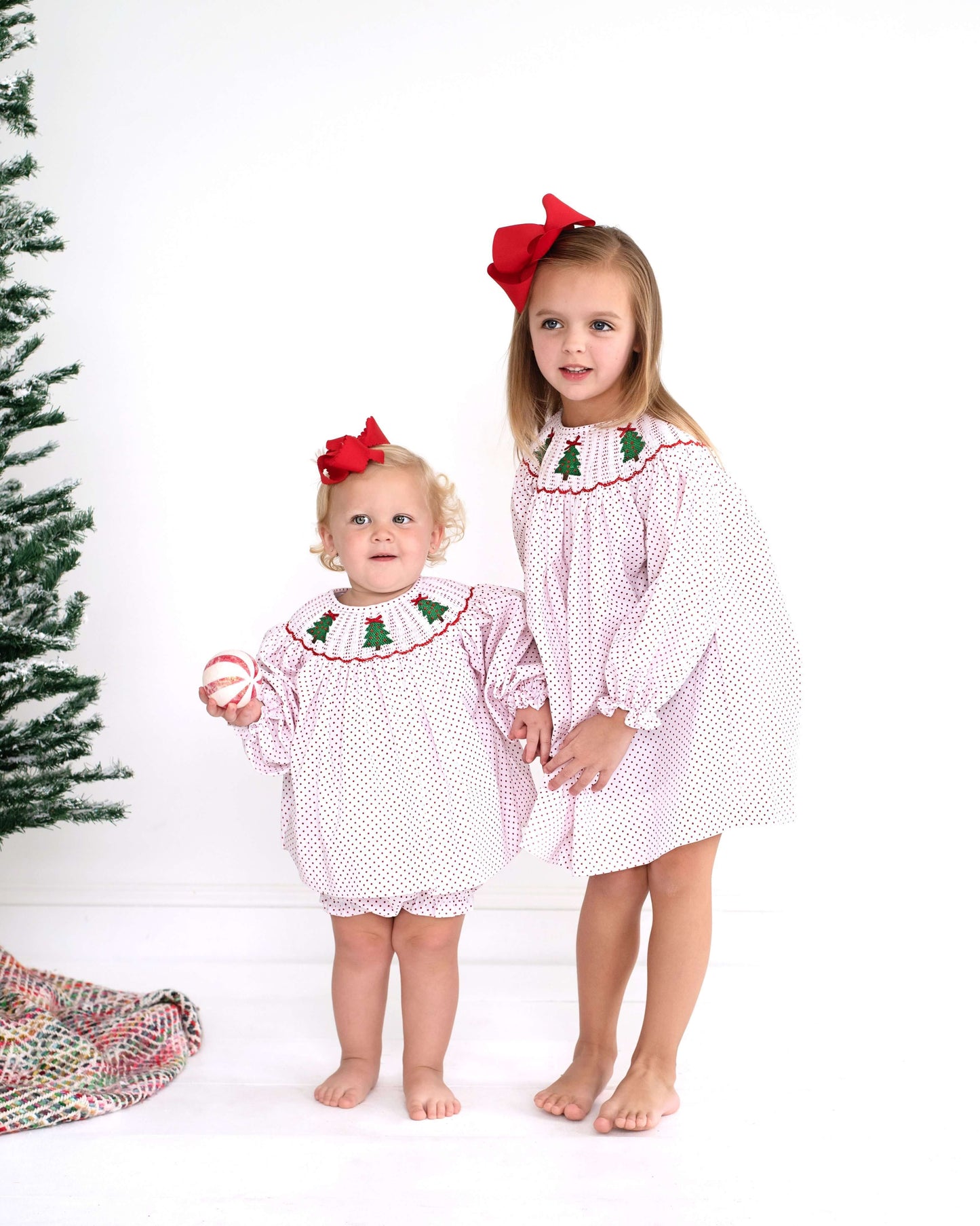 Two children in matching smocked Christmas-themed outfits standing beside a Christmas tree.
