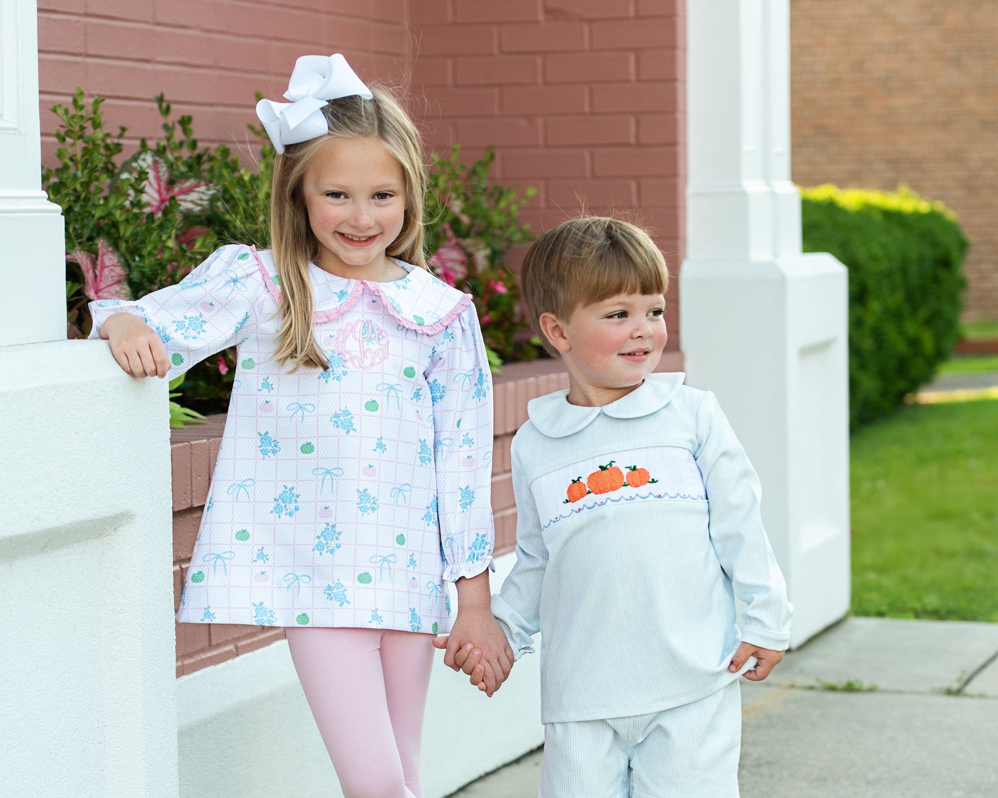 Two children in pumpkin outfits standing on a porch with a brick wall and greenery in the background 1