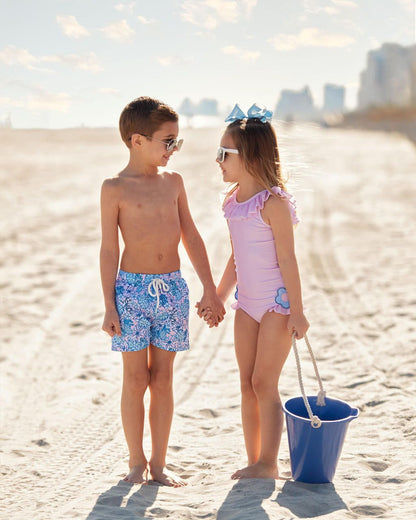 Two children on a beach, one in floral shorts and the other in a pink swimsuit with a blue bucket.