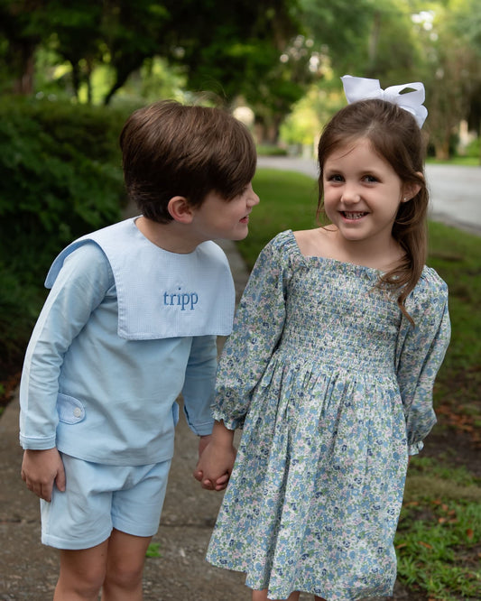 Two children outdoors holding hands, wearing blue and floral printed outfits. The boy wears a light blue long-sleeve top and shorts with a white bib, and the girl wears a light blue floral long-sleeve dress with a white bow in her hair.