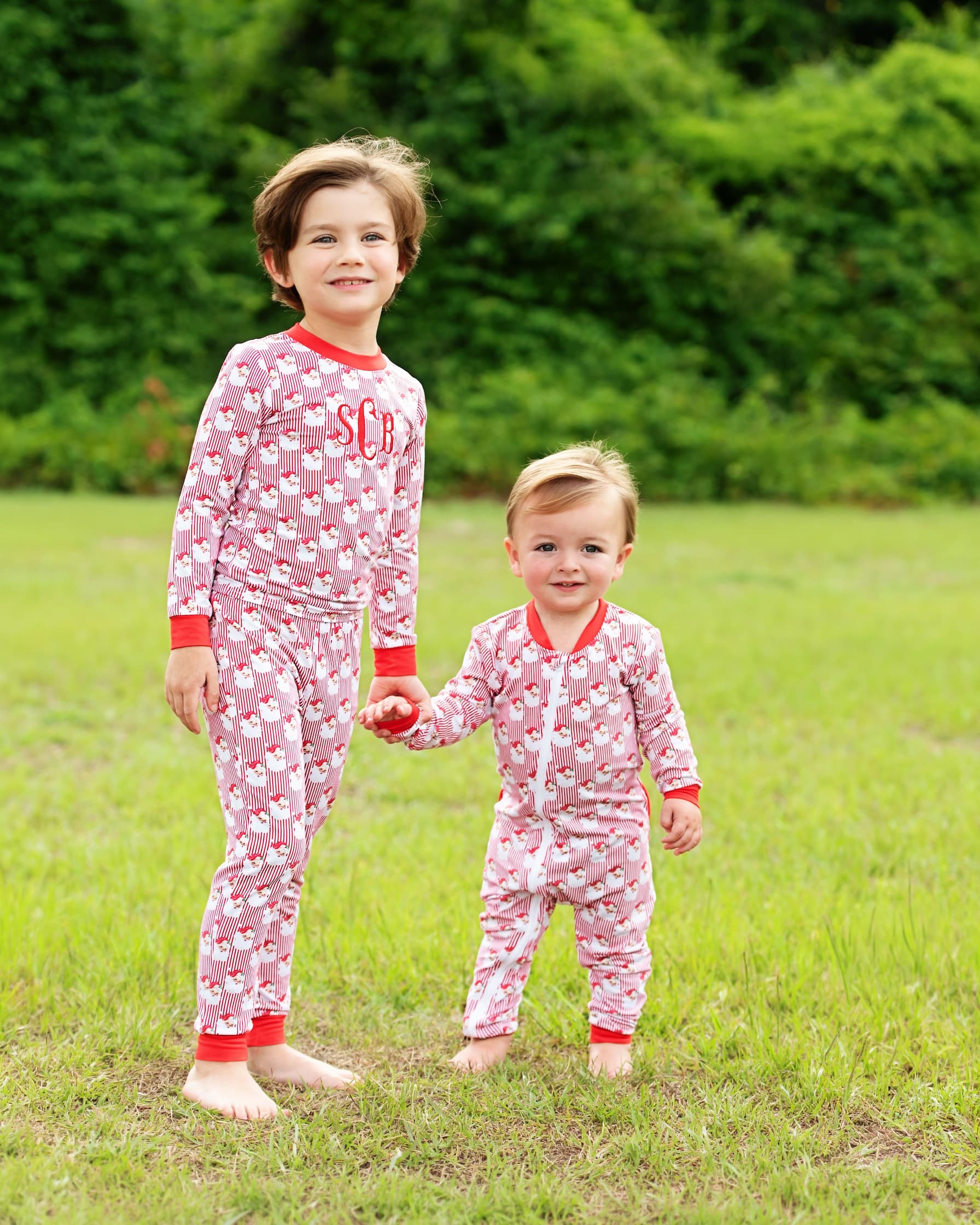 Two children standing barefoot on a grassy lawn outdoors, wearing matching pink and white patterned pajamas with red trim. The older child has 'SC' printed on the chest, and the younger child wears a front zipper.