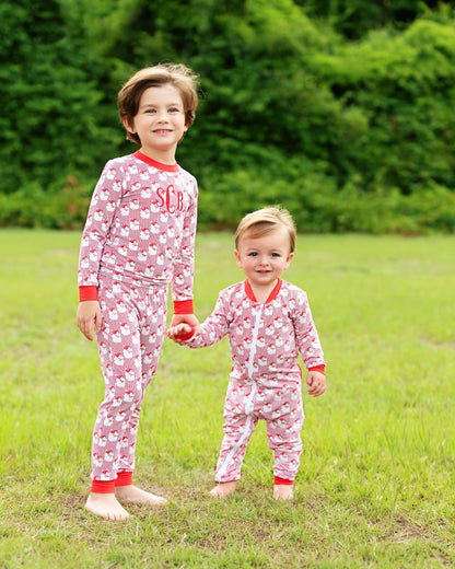 Two children standing barefoot on a grassy lawn outdoors, wearing matching pink and white patterned pajamas with red trim. The older child has 'SC' printed on the chest, and the younger child wears a front zipper.