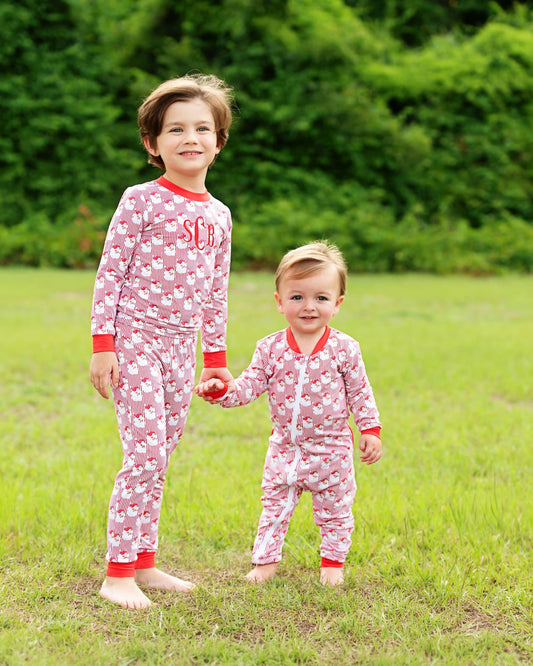 Two children standing barefoot on a grassy lawn outdoors, wearing matching pink and white patterned pajamas with red trim. The older child has 'SC' printed on the chest, and the younger child wears a front zipper.