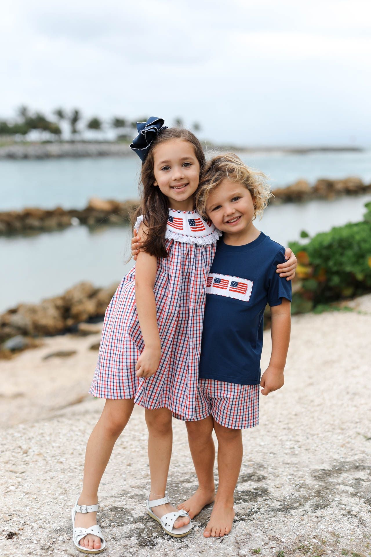 Two children standing on a pebbly beach with water and greenery in the background.