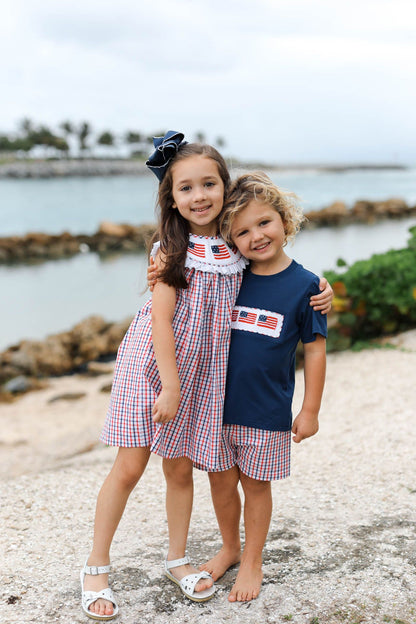 Two children standing on a pebbly beach with water and greenery in the background.