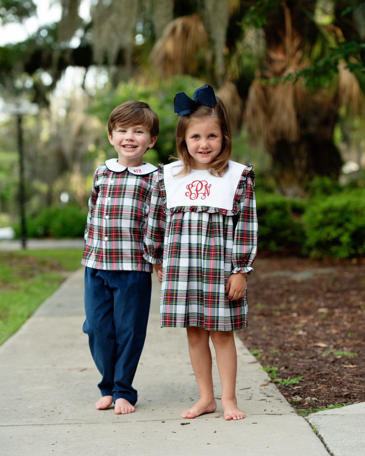 Two children standing on a sidewalk outdoors, wearing matching plaid outfits. 