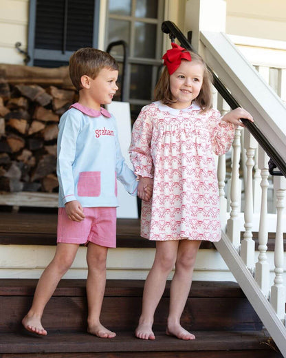 Two children standing on a staircase, one in a light blue shirt and red shorts, the other in a patterned dress with a red bow.