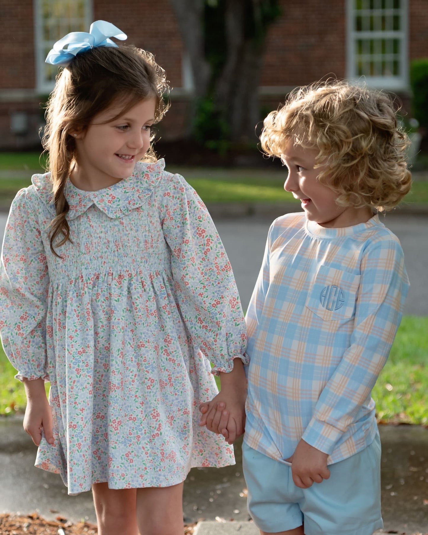 Two children standing outdoors and holding hands. The girl wears a light blue floral long-sleeve dress with a white collar and a blue bow in her hair. The boy wears a plaid shirt and blue shorts