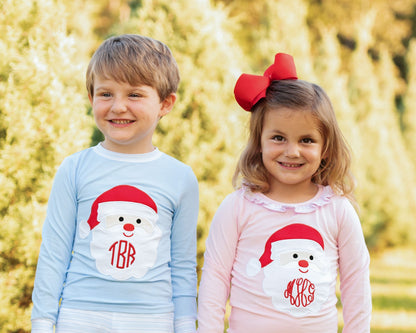 Two children standing outdoors in front of a blurred yellow-green background, wearing matching long-sleeve shirts with a Santa Claus graphic. The child on the left wears a light blue shirt with a white Santa graphic and red monogram '1BR', and the child on the right wears a light pink shirt with a white Santa graphic and red monogram 'MRO'.
