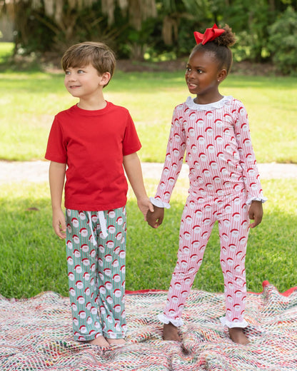 Two children standing outdoors on a blanket in a grassy yard, wearing matching holiday-themed pajamas. The boy wears a red short-sleeve shirt with light green pants printed with a red and white pattern.