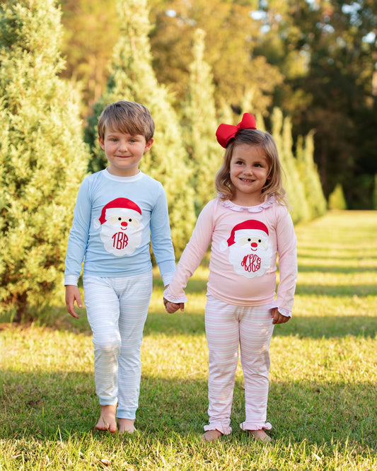 Two children standing outdoors on grass holding hands, wearing matching long-sleeve pajama sets with a snowman graphic and monogram. The boy wears a light blue set, and the girl wears a light pink set.