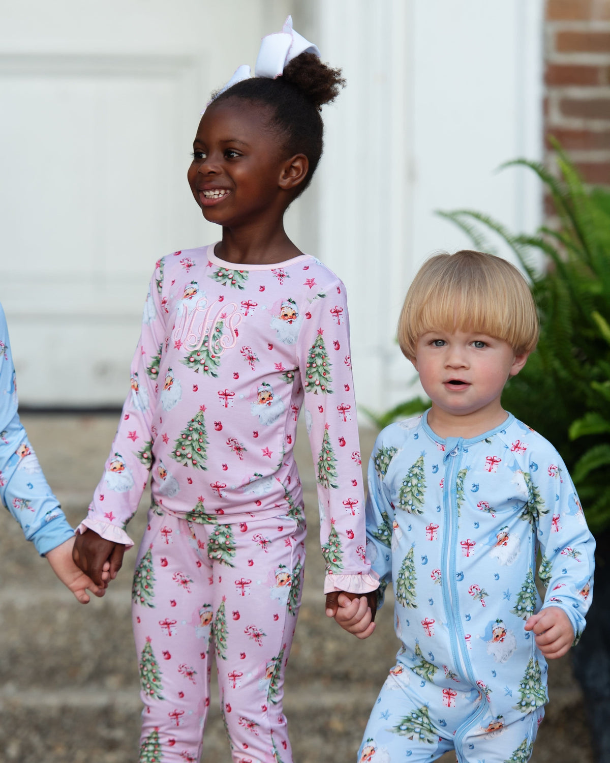 Two children wearing matching holiday-themed pajamas, with the girl in a pink long-sleeve pajama set and the boy in a light blue one-piece, both printed with Christmas trees and ornaments, standing outdoors and holding hands.