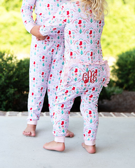 Two children wearing matching long-sleeve pajama sets with a colorful winter holiday print, standing barefoot outdoors on a concrete surface with green bushes in the background.