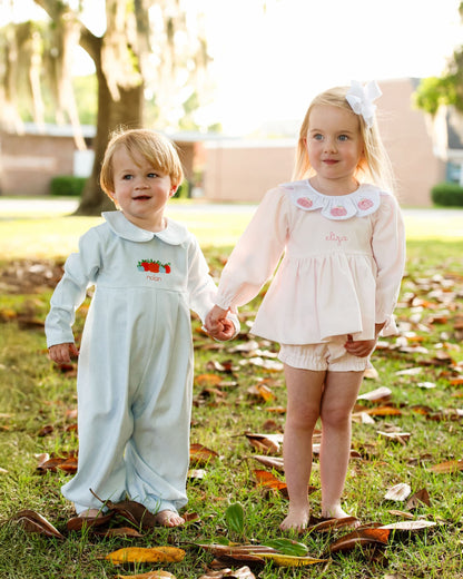 Two children with pumpkin outfits holding hands on a grassy area with fallen leaves.