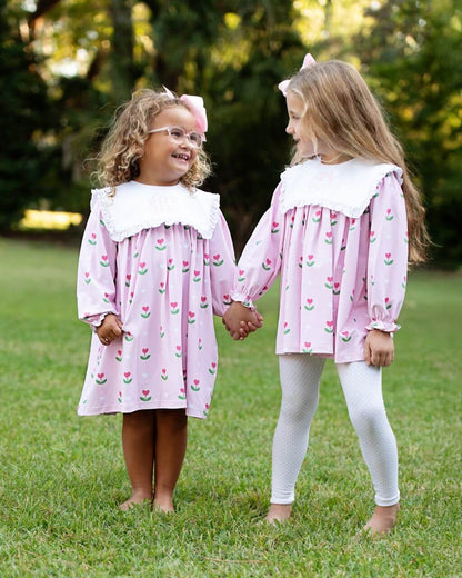 Two girls in matching pink dresses with white ruffled collars standing on grass.