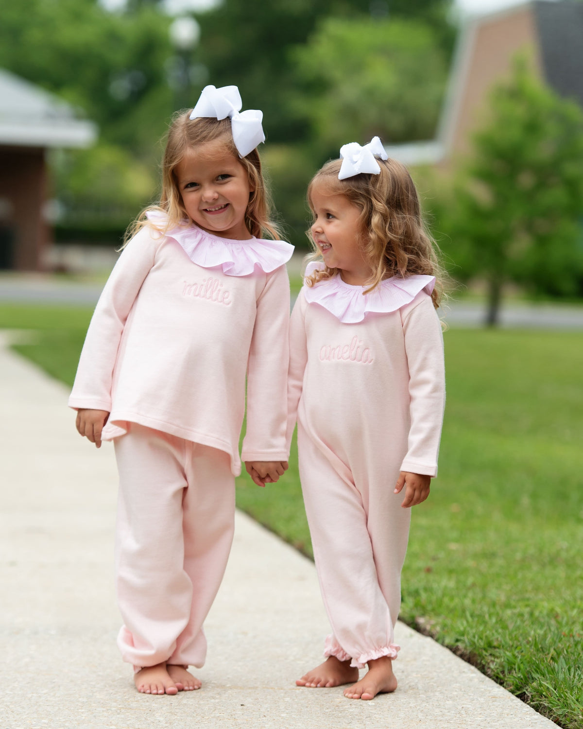 Two girls wearing matching pink long-sleeve pajamas with ruffled collars and white bows in their hair, standing barefoot on a concrete path in a backyard.