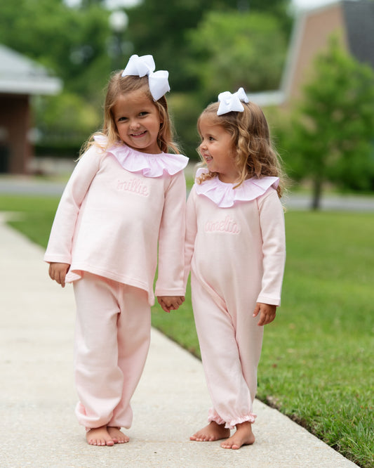 Two girls wearing matching pink long-sleeve pajamas with ruffled collars and white bows in their hair, standing barefoot on a concrete path in a backyard.