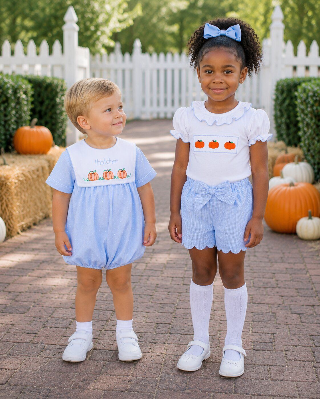 Two kids in matching outfits with pumpkin designs standing on a path with pumpkins in the background.