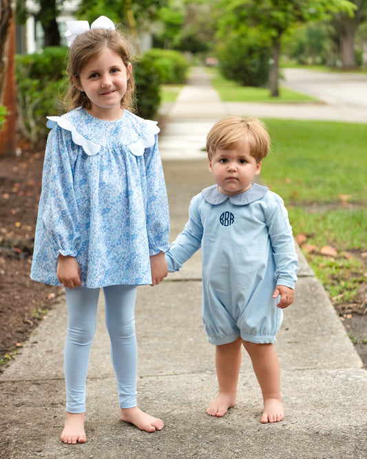 Two toddlers standing on a concrete sidewalk outdoors, holding hands. The girl wears a light blue floral long-sleeve tunic with a white ruffled collar and matching light blue leggings.