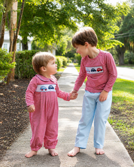 Two toddlers standing on a paved path outdoors, holding hands. Both are wearing matching pink and red striped long-sleeve pajamas with a small car graphic on the chest, paired with one child in light blue pants and the other in pink pants.