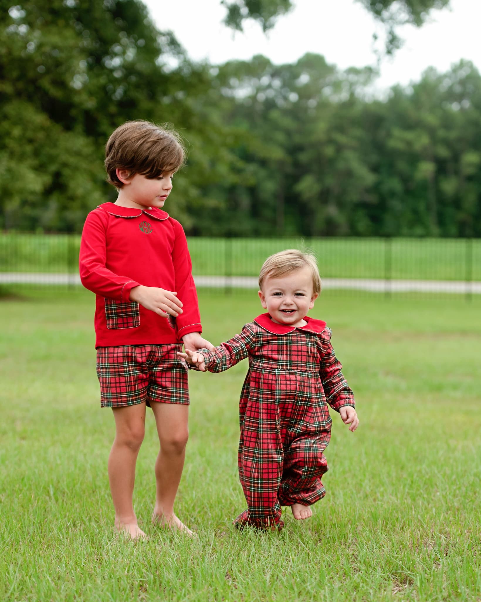 Two young boys outdoors on a grassy field wearing matching red and green plaid holiday-themed outfits, with trees in the background.