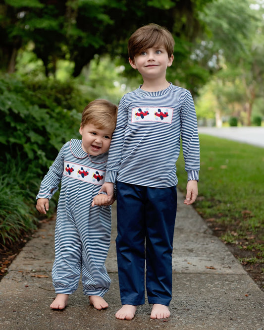 Two young boys standing barefoot on a sidewalk outdoors, wearing matching long-sleeve striped outfits with red airplane motifs and dark navy pants.