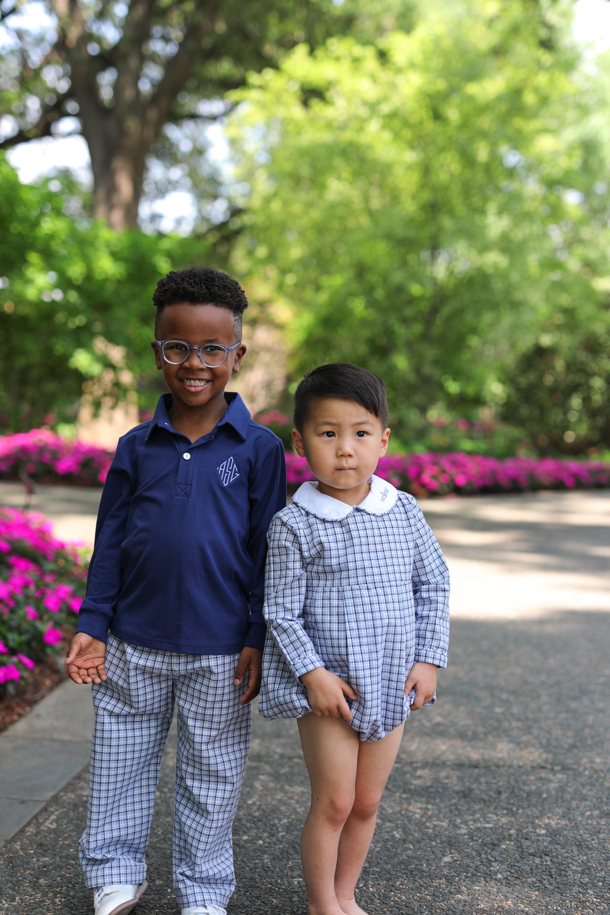 Two young boys standing outdoors on a paved path in a garden. The older boy wears a navy blue long-sleeve polo shirt with a small white monogram on the chest and matching blue-and-white checkered pants.