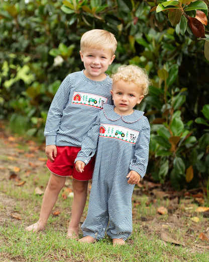 Two young boys standing outdoors on grass in front of dense green foliage, wearing matching long-sleeve striped pajamas with a smocked tractor design on the chest. The older boy also wears red shorts.