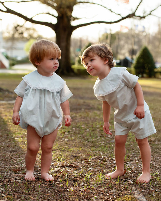Two young children in light-colored outfits standing outdoors with trees in the background.