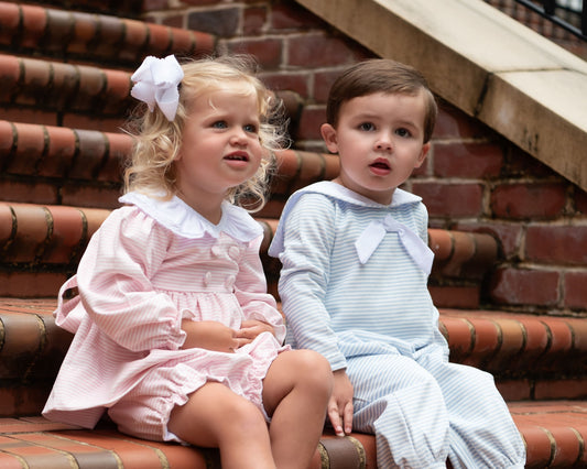 Two young children sitting on brick stairs outdoors. The girl wears a light pink pinstriped dress with a white ruffled collar and a white bow in her hair.