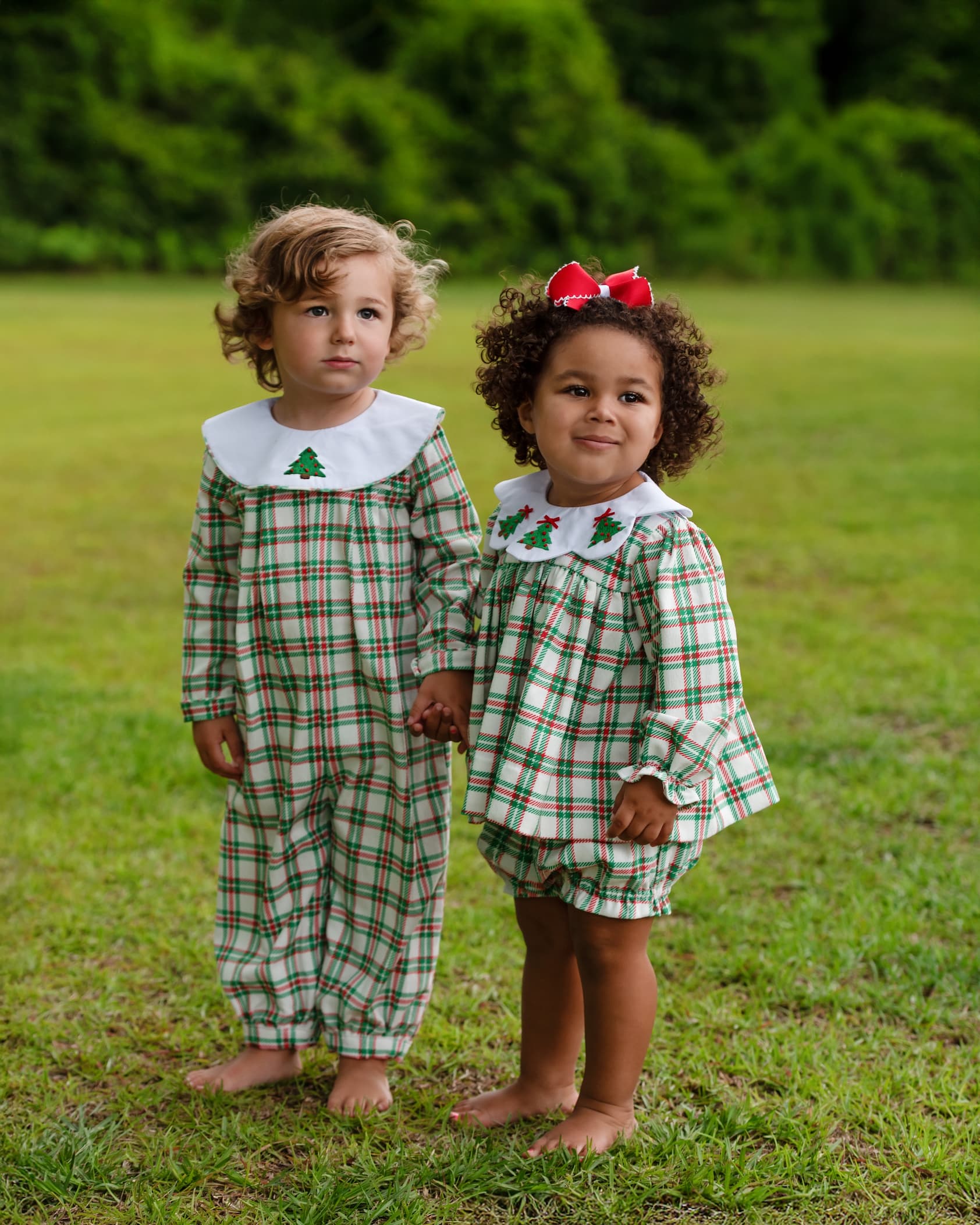 Two young children standing barefoot on a grassy lawn outdoors, wearing matching long-sleeve plaid outfits in green, white, and red with white Peter Pan collars and small green tree motifs.