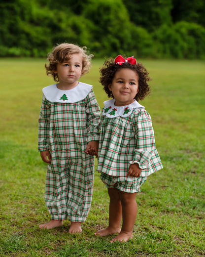 Two young children standing barefoot on a grassy lawn outdoors, wearing matching long-sleeve plaid outfits in green, white, and red with white Peter Pan collars and small green tree motifs.