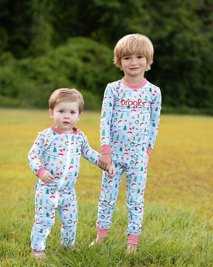 Two young children standing in a grassy field outdoors, wearing matching long-sleeve pajamas with a light blue background and a Christmas print. The pajamas feature a multicolor holiday pattern with red, green, and white motifs, and the word 'brooks' is printed in red on the chest of the taller child's pajama.