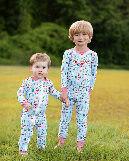 Two young children standing in a grassy field outdoors, wearing matching long-sleeve pajamas with a light blue background and a Christmas print. The pajamas feature a multicolor holiday pattern with red, green, and white motifs, and the word 'brooks' is printed in red on the chest of the taller child's pajama.