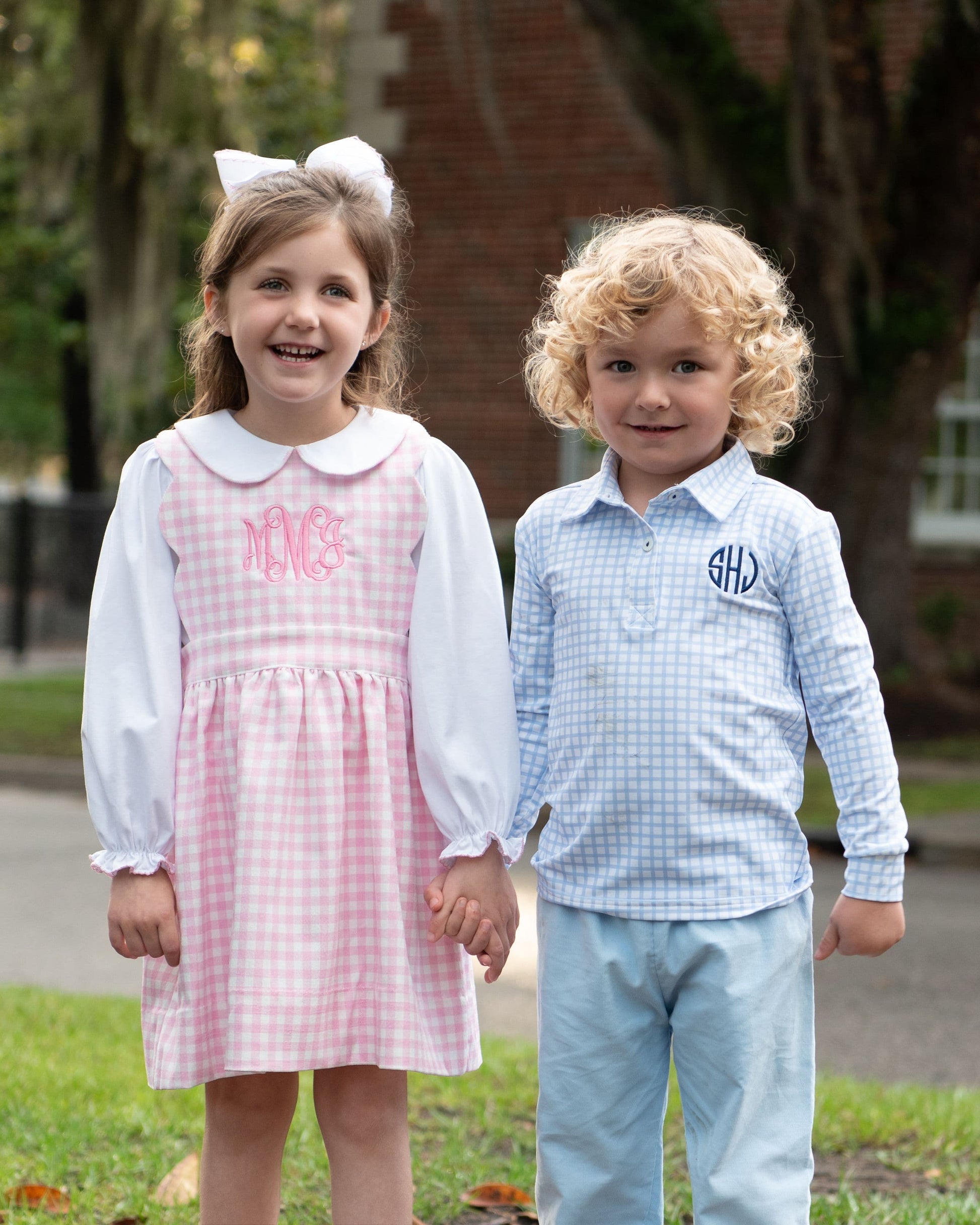 Two young children standing outdoors holding hands. The girl wears a pink and white gingham dress with long white sleeves and a white Peter Pan collar, with pink monogram lettering on the chest.