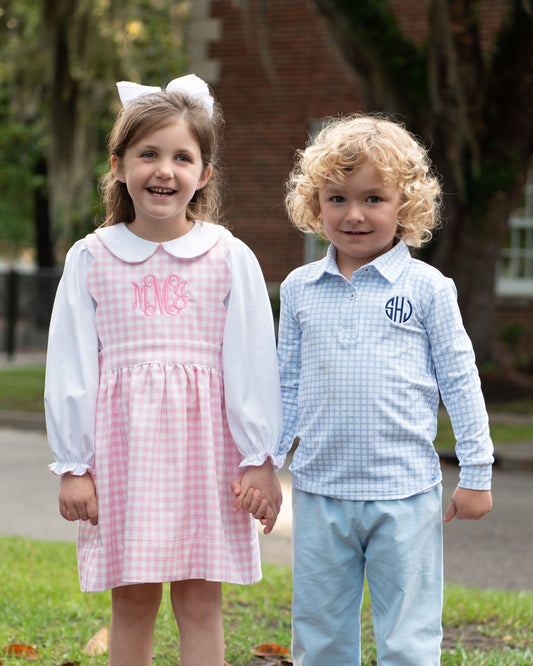 Two young children standing outdoors holding hands. The girl wears a pink and white gingham dress with long white sleeves and a white Peter Pan collar, with pink monogram lettering on the chest.