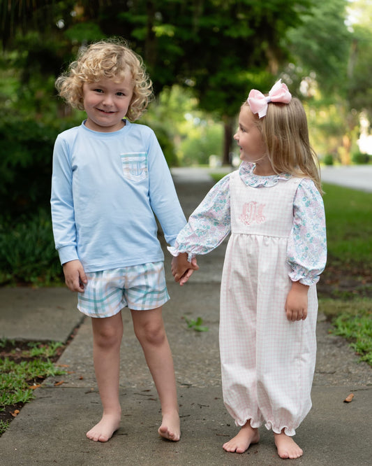 Two young children standing outdoors on a paved path holding hands. The boy wears a light blue long-sleeve shirt and plaid shorts, and the girl wears a white gingham bonnet dress with pastel floral sleeves and a pink bow in her hair.