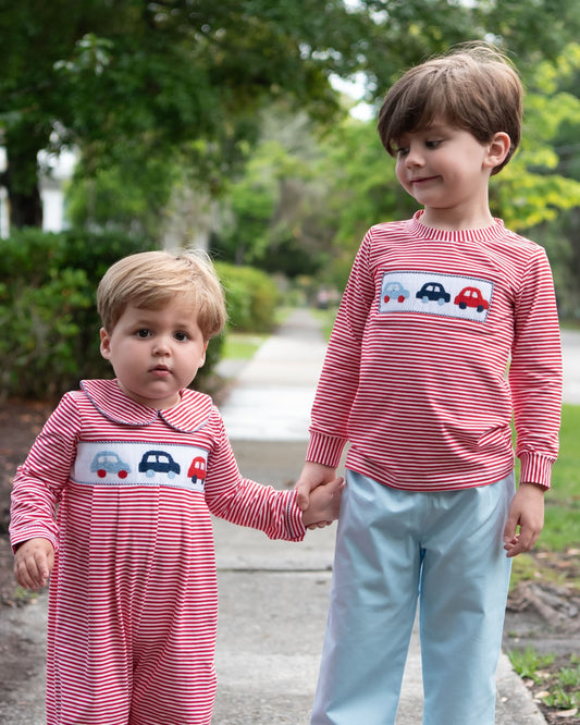 Two young children standing outdoors on a paved path, holding hands. The child on the left wears a red and white striped footless sleeper with a car graphic on the chest, and the child on the right wears a matching red and white striped long-sleeve shirt with light blue pants.