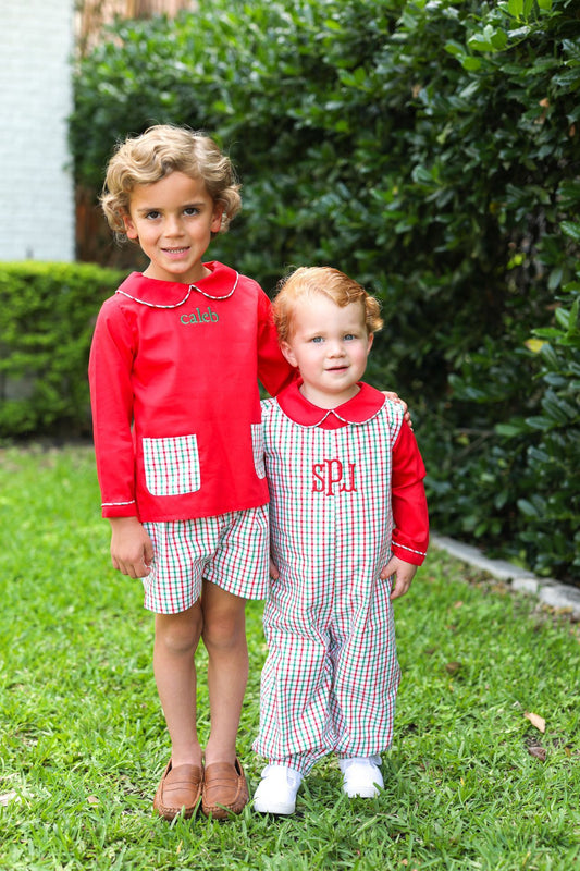 Two young children standing outdoors on grass in front of a hedge, wearing matching red and checkered long-sleeve outfits. The older child wears shorts and the younger child wears a one-piece overall, both with red collars and cuffs.