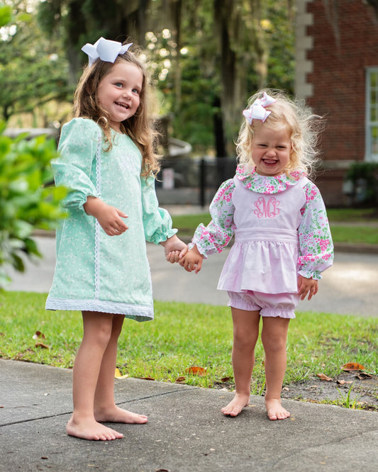 Two young girls holding hands on a sidewalk outdoors, wearing matching floral and solid long-sleeve dresses with bows in their hair. The girl on the left wears a mint green dress, and the girl on the right wears a light pink dress with a floral collar and sleeves.