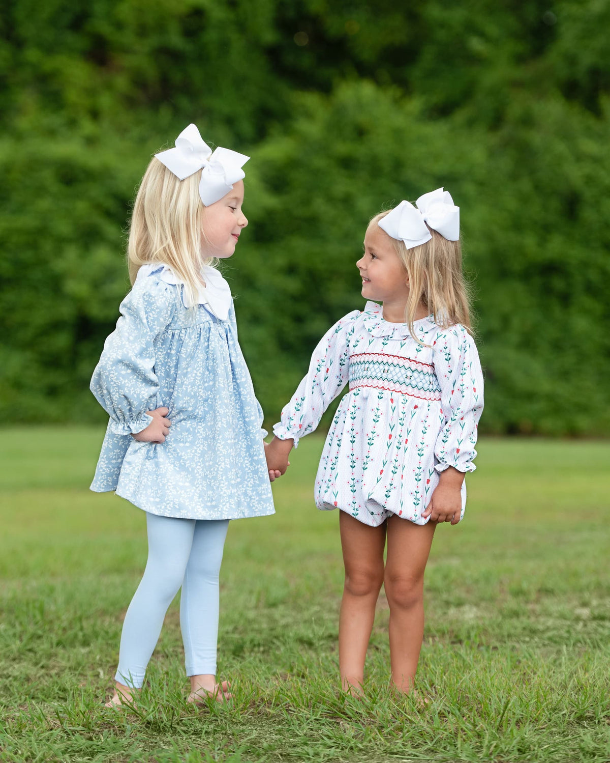 Two young girls holding hands standing on the grass wearing floral outfits