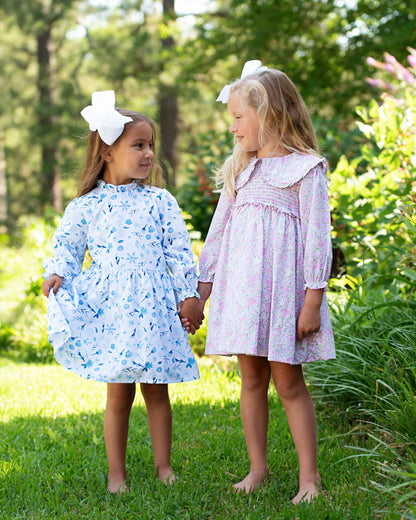 Two young girls in floral dresses standing in a grassy area with trees in the background
