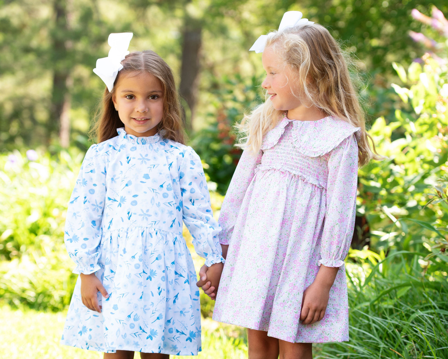 Two young girls in floral dresses standing outdoors with greenery in the background