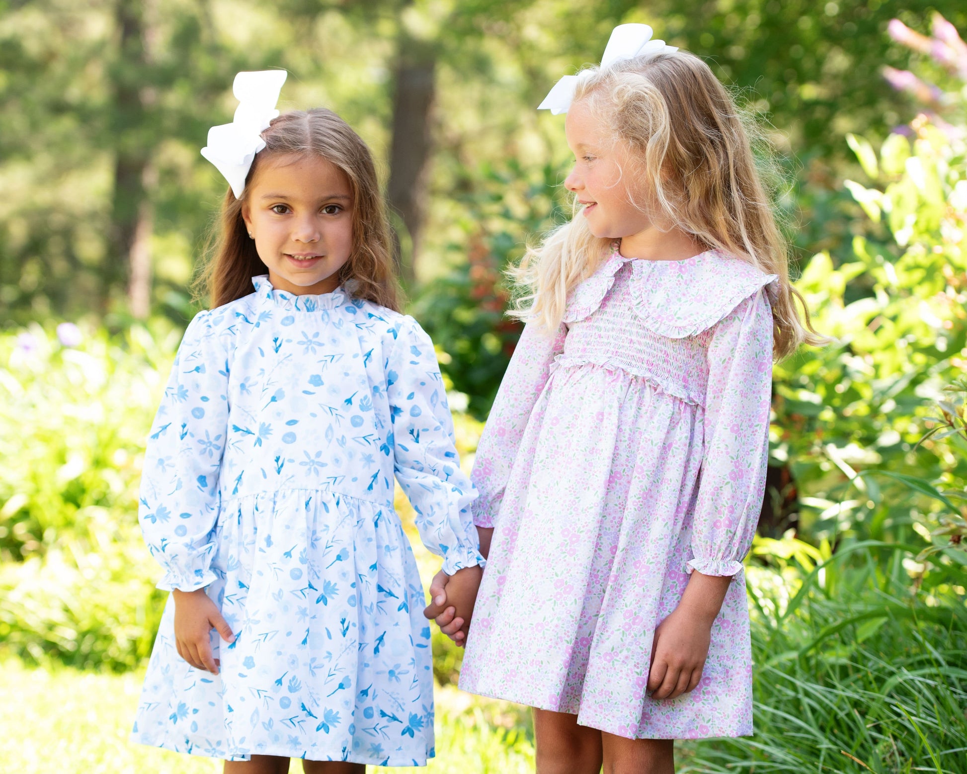 Two young girls in floral dresses standing outdoors with greenery in the background