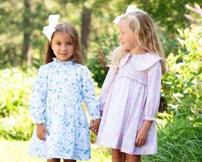 Two young girls in floral dresses standing outdoors with greenery in the background