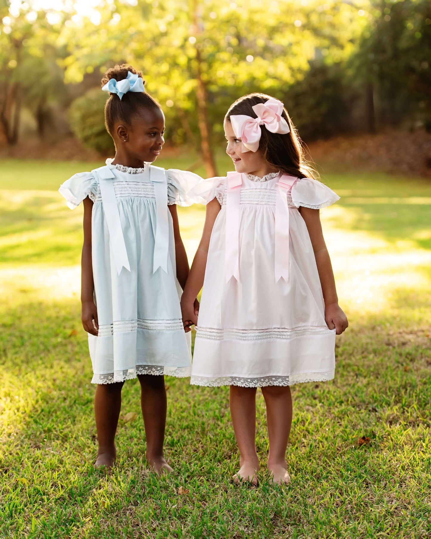 Two young girls in light dresses standing in a grassy field with trees in the background.