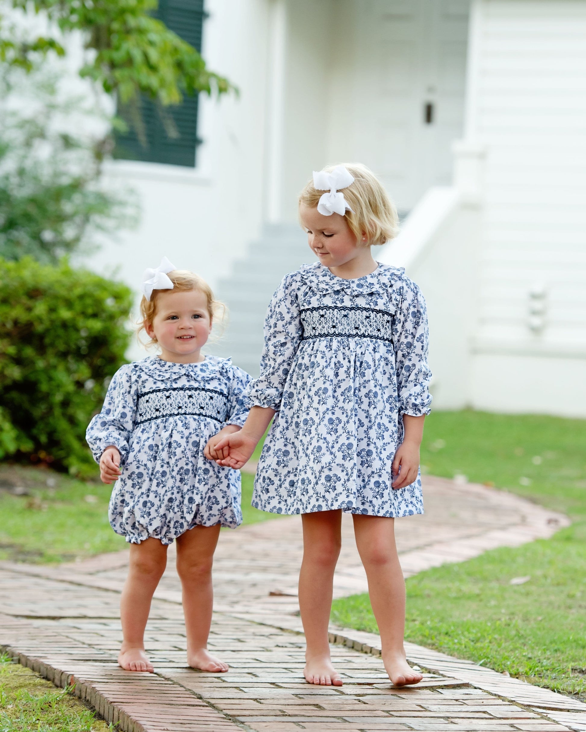 Two young girls in matching blue floral outfits standing on a wooden path outdoors.