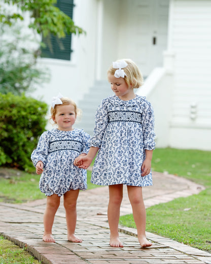 Two young girls in matching blue floral outfits standing on a wooden path outdoors.